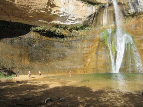 A serene waterfall cascades into a pool, with people enjoying the natural beauty in a sunlit canyon.