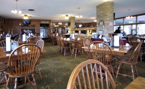 A cozy restaurant interior with wooden tables, chairs, and a view of the kitchen area in the background.