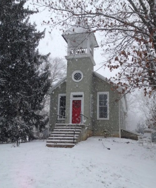 A quaint green house with a red door, surrounded by snow and trees, during a snowy winter day.
