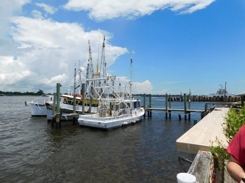 Fishing boats docked at a pier on a sunny day, with blue skies and fluffy clouds above.