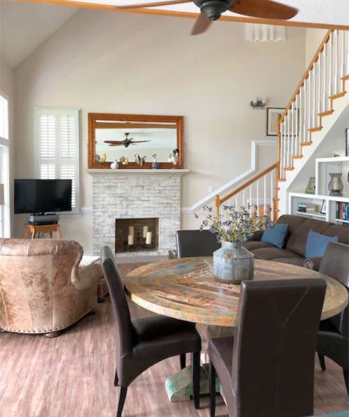 Cozy living room with a wooden dining table, leather chair, stone fireplace, and large windows.