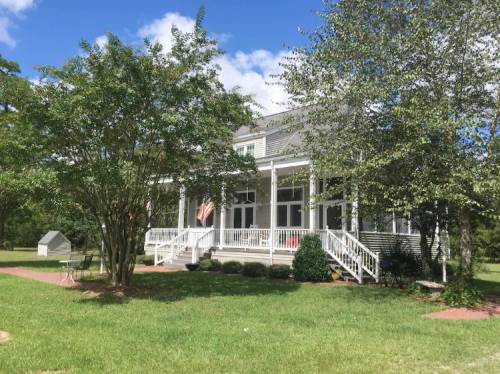 A large, two-story house with a porch, surrounded by trees and a grassy yard, under a blue sky with clouds.