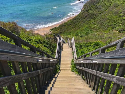 A wooden staircase leads down to a sandy beach, surrounded by lush greenery and ocean waves in the background.