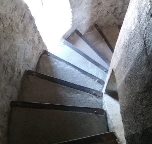 A narrow stone staircase with metal railings, leading downwards, illuminated by light from a window above.