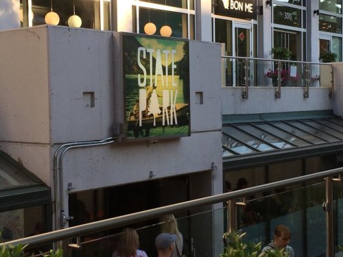 Sign for "STATE PARK" displayed on a building exterior, with people seated nearby and plants in view.
