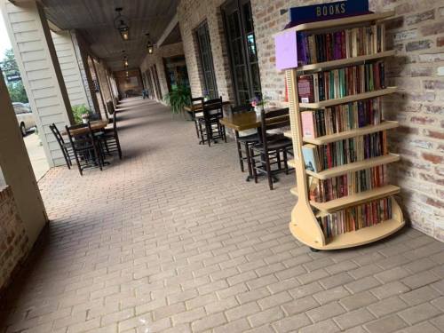 A brick walkway lined with tables and chairs, featuring a wooden bookshelf filled with books.
