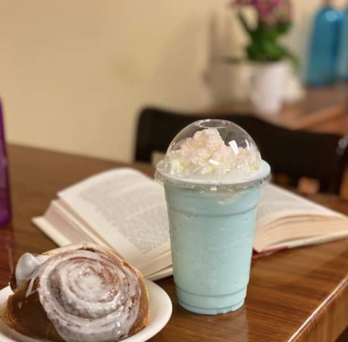 A blue milkshake topped with whipped cream and a cinnamon roll on a wooden table, with a book in the background.