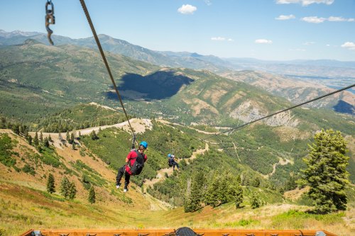 Two people ziplining over a lush green mountain landscape with distant hills and a clear blue sky.