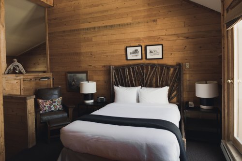 Cozy wooden bedroom with a double bed, two lamps, a chair, and framed pictures on the wall. Natural light from a window.