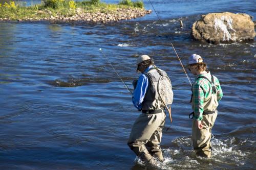 Two anglers wade through a river, fishing rods in hand, surrounded by water and rocky banks.