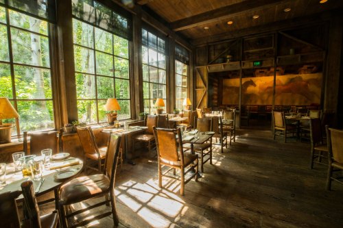 Cozy restaurant interior with wooden tables and chairs, large windows, and natural light streaming in from outside.