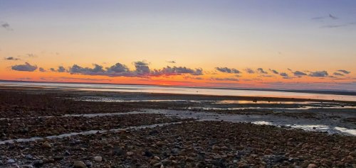 A serene sunset over a calm beach, with colorful clouds reflecting on the water and rocky shore in the foreground.