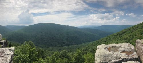 A panoramic view of lush green mountains under a partly cloudy sky. Rocky foreground with expansive valleys in the distance.