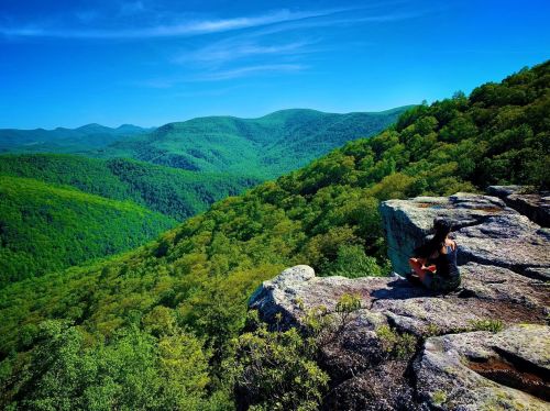 A person sits on a rocky ledge, overlooking a lush green valley and rolling hills under a clear blue sky.