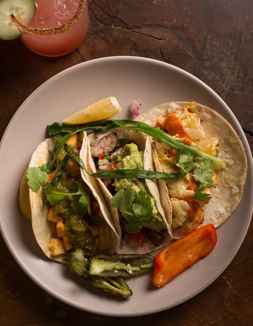 Three tacos filled with various toppings, garnished with cilantro, served on a plate with lemon and peppers.