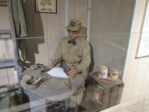A lifelike mannequin in a workshop, wearing a uniform and bow tie, working on a piece of paper with tools around.