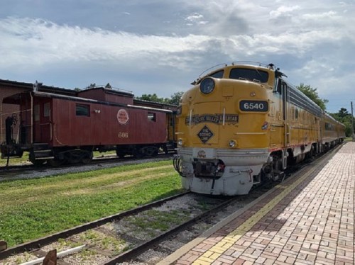 A vintage yellow train engine parked beside a red caboose on a sunny day, with green grass and a brick path nearby.