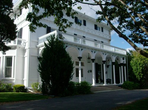 A large white house with a porch, arched columns, and lush greenery surrounding it on a sunny day.
