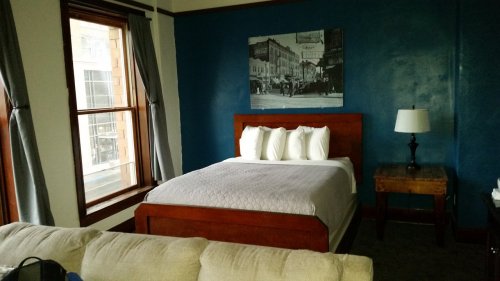 Cozy hotel room featuring a bed, wooden furniture, a lamp, and a blue accent wall with a vintage photo.