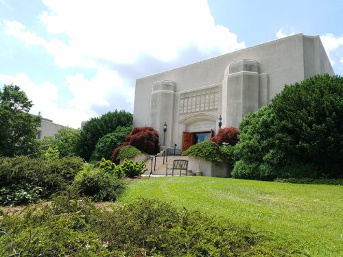 A modern building with a decorative entrance, surrounded by lush greenery and colorful shrubs under a partly cloudy sky.