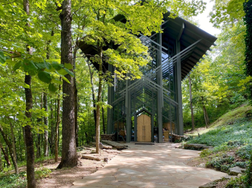 A modern glass chapel surrounded by lush green trees and a stone pathway leading to its entrance.