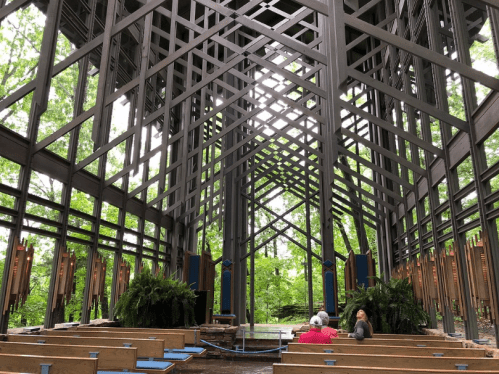 Interior of a modern, open chapel with wooden beams and greenery visible through large windows, two people seated.