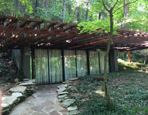 A modern house with a wooden overhang, surrounded by trees and greenery, featuring large glass windows.