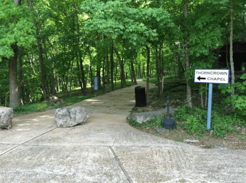 Pathway through a green forest with a sign pointing left to Thorncrown Chapel. Large rocks line the path.