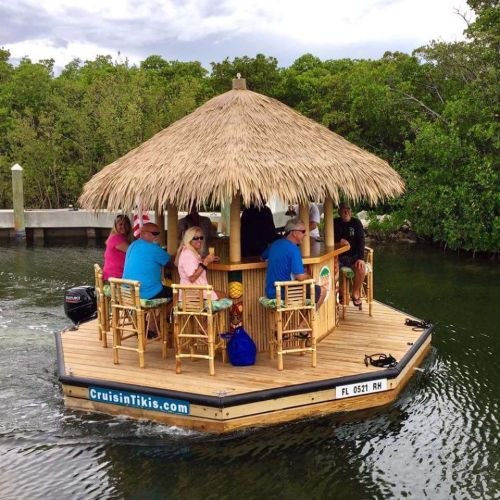 A tiki boat with a thatched roof, featuring people enjoying drinks and sitting at a bar, surrounded by water and greenery.