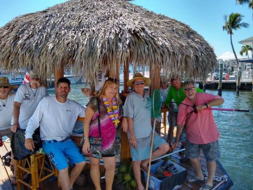 A group of people smiling under a thatched roof by the water, enjoying a sunny day together.