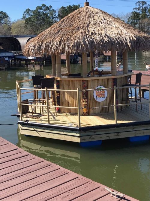 A tiki bar on a floating platform, surrounded by water, with a thatched roof and wooden deck.