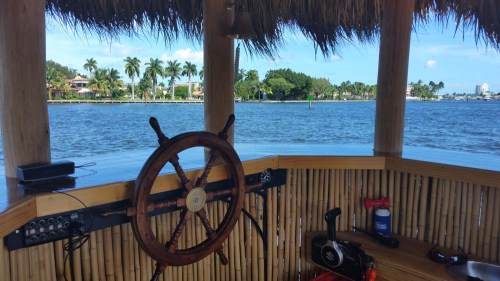 A wooden steering wheel in a tiki hut overlooking a calm waterway with palm trees and houses in the background.