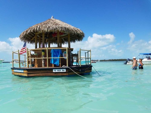 A floating tiki bar with a thatched roof in clear blue water, with people swimming nearby and boats in the background.