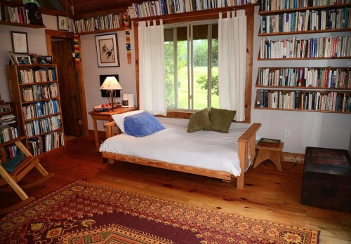 Cozy room with a wooden daybed, bookshelves filled with books, and a view of greenery through a large window.