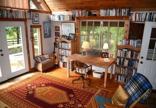 Cozy home office with a wooden desk, bookshelves, guitar, and large windows overlooking a green landscape.