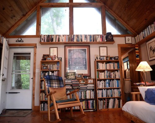 Cozy room with wooden walls, large windows, bookshelves filled with books, a rocking chair, and soft lighting.