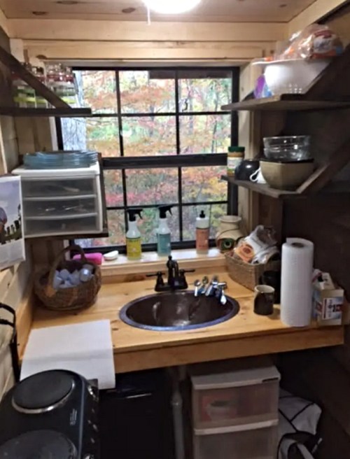 Cozy kitchen with wooden shelves, a sink, and various kitchen items, overlooking a colorful autumn landscape through a window.