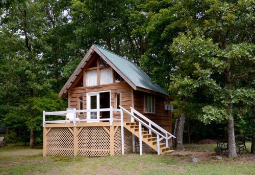 A charming wooden cabin with a green metal roof, surrounded by trees and featuring a front porch and steps.