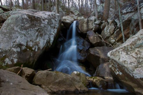 A serene waterfall cascades over large rocks in a wooded area, surrounded by trees and natural scenery.