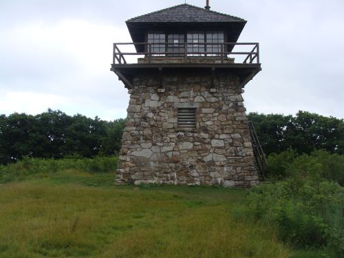 A stone watchtower with a wooden balcony, surrounded by grassy fields and trees under a cloudy sky.