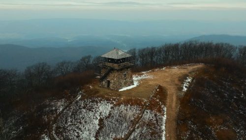 A stone lookout tower on a snowy hilltop, surrounded by bare trees and distant mountains under a cloudy sky.