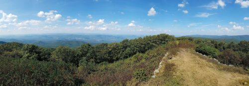 A panoramic view of rolling hills and valleys under a blue sky with scattered clouds, surrounded by lush greenery.