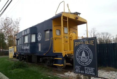 A blue and yellow B&O caboose on display next to a sign, set against a backdrop of trees and a clear sky.