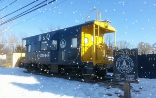 A blue and yellow train caboose sits in the snow, with a sign nearby reading "The Royal Blue." Snowflakes are falling.