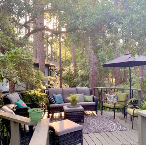 Cozy outdoor seating area with a sofa, chairs, and a table, surrounded by trees and soft sunlight.