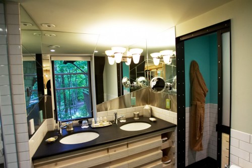 Modern bathroom with double sinks, large mirrors, and a window overlooking greenery. Warm lighting and stylish decor.