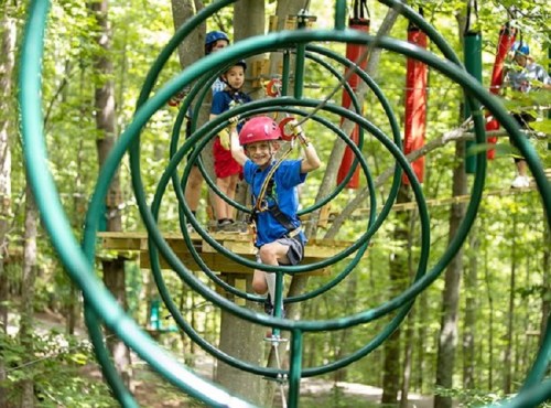 A child in a helmet navigates a circular rope course in a forest, with another child visible in the background.