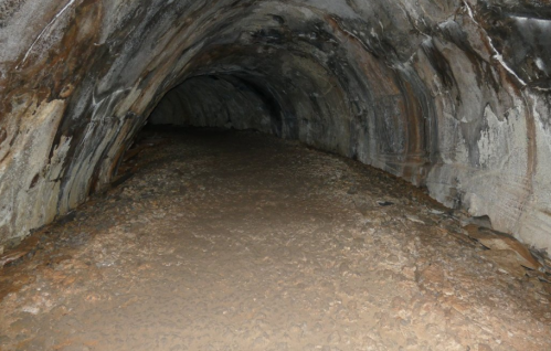 A dimly lit cave interior with a sandy floor and curved rock walls, creating a tunnel-like appearance.