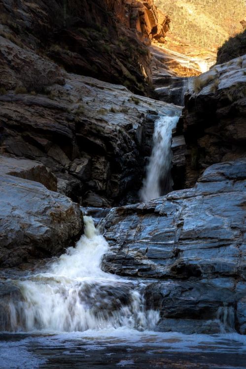 A cascading waterfall flows over rocky terrain, surrounded by steep cliffs and illuminated by warm sunlight.