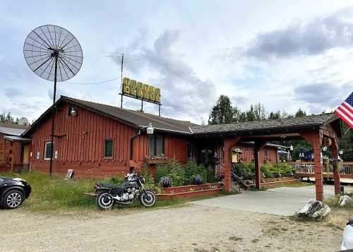 A rustic wooden building with a satellite dish, motorcycle, and an American flag outside, set in a grassy area.